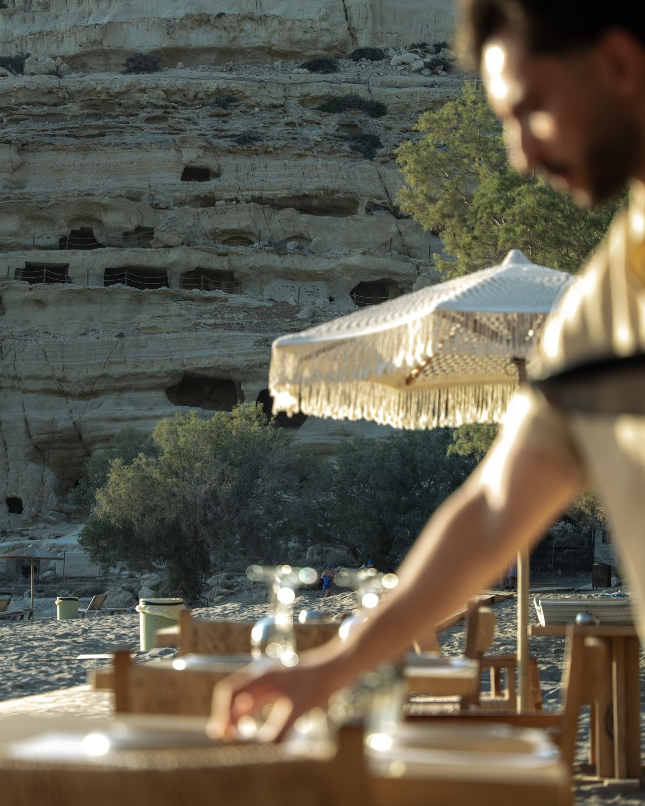 Waiter setting a beach table beneath the caves of Matala